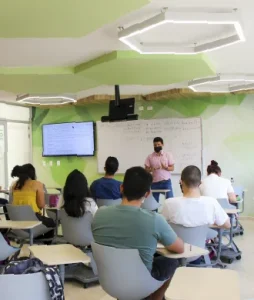 Plano general donde se ve a un grupo de estudiantes recibiendo clase y al profesor explicando en el tablero