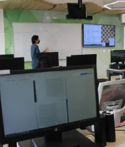 Primer plano de la pantalla de un computador y un estudiante en el fondo explicando frente al tablero